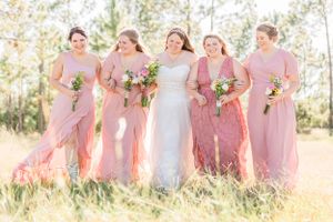Wedding party of bridesmaids in pink dresses poses together in sunlit field.