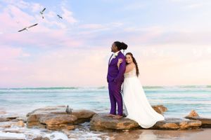 A couple stands on coastal rocks with seabirds flying overhead at sunset.