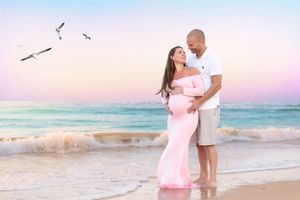 Expecting couple embraces on beach at sunset with seabirds flying overhead.