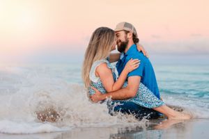 A couple splashing in ocean waves during sunset with warm pink sky in background.