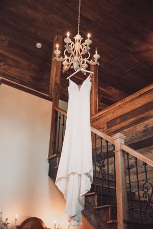 An elegant white wedding dress hanging from an ornate chandelier against a wooden ceiling and staircase.