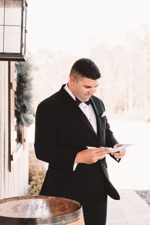 A groom in black tuxedo reading a letter while standing next to a rustic wine barrel.