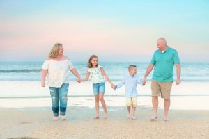 Family holding hands while walking together on beach at sunset in pastel colored outfits.
