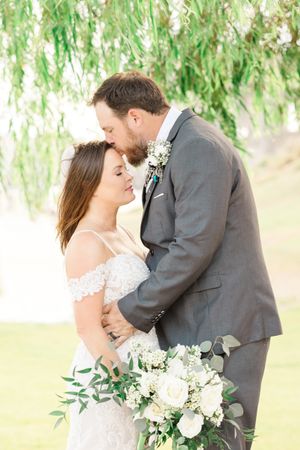 Wedding couple sharing tender moment under willow tree with white floral bouquet.