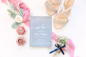 Wedding details flatlay with blue invitation, pink accents, and delicate sandals.