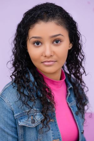 Portrait against purple backdrop wearing pink top and denim jacket.