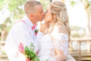 Wedding couple sharing romantic kiss in white attire with pink floral bouquet.