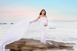 Maternity photo of woman in white dress with flowing fabric on rocky beach overlook.