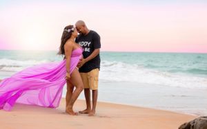 Romantic couple embraces on beach at sunset with flowing pink dress against ocean waves.