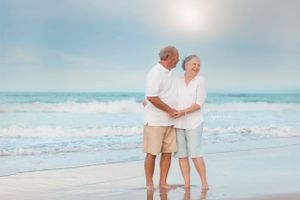 Elderly couple in white clothing embracing while standing on a tranquil beach at sunset.