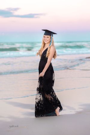 Graduate in black lace dress and cap poses on beach during sunset.