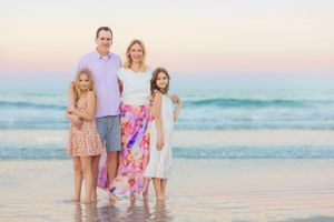 Family of four standing together on beach at sunset in coordinated pastel outfits.