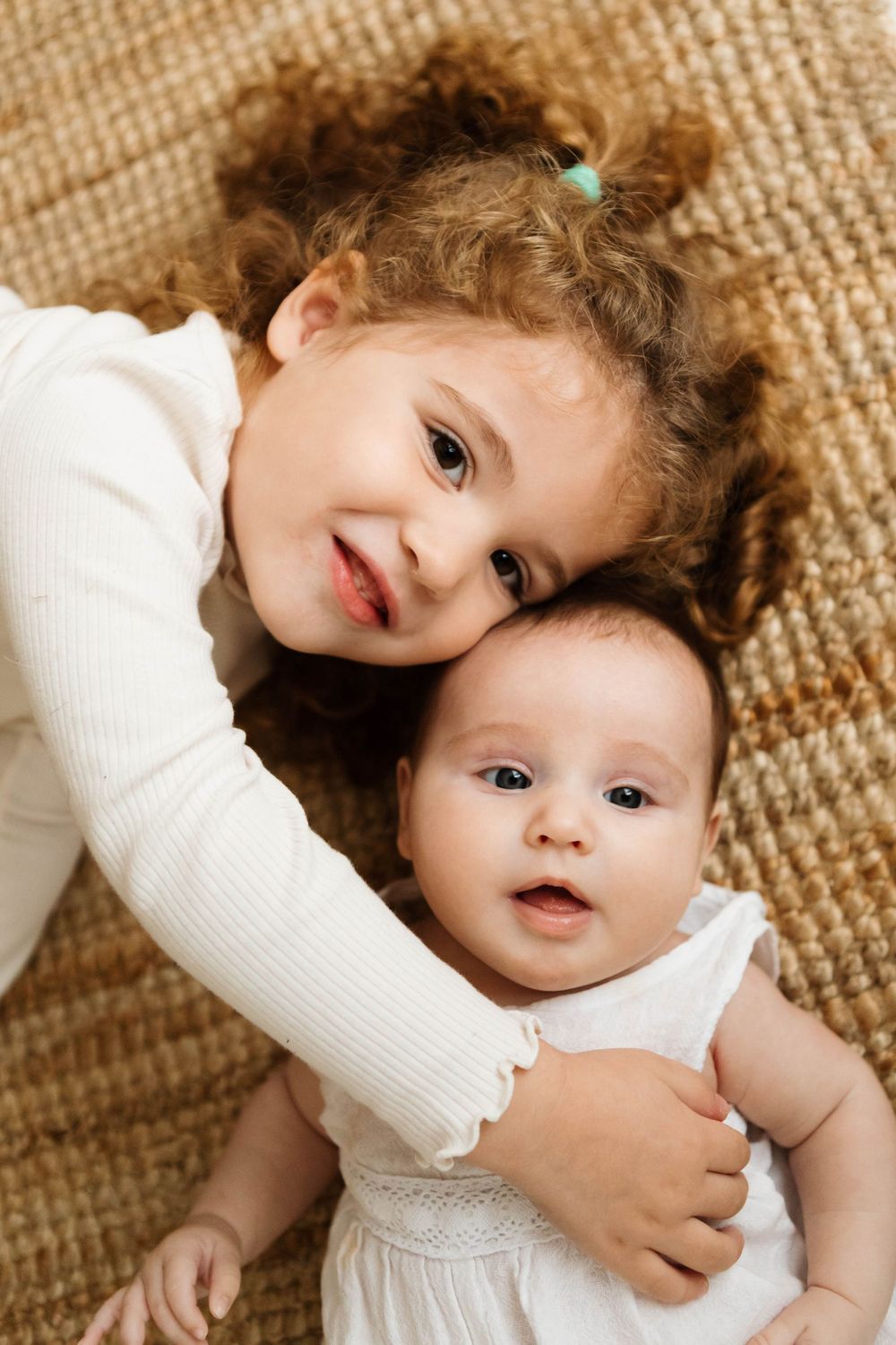 Two young children wearing white outfits lie close together on a textured beige carpet sharing a sweet moment in Cambridge Ontario