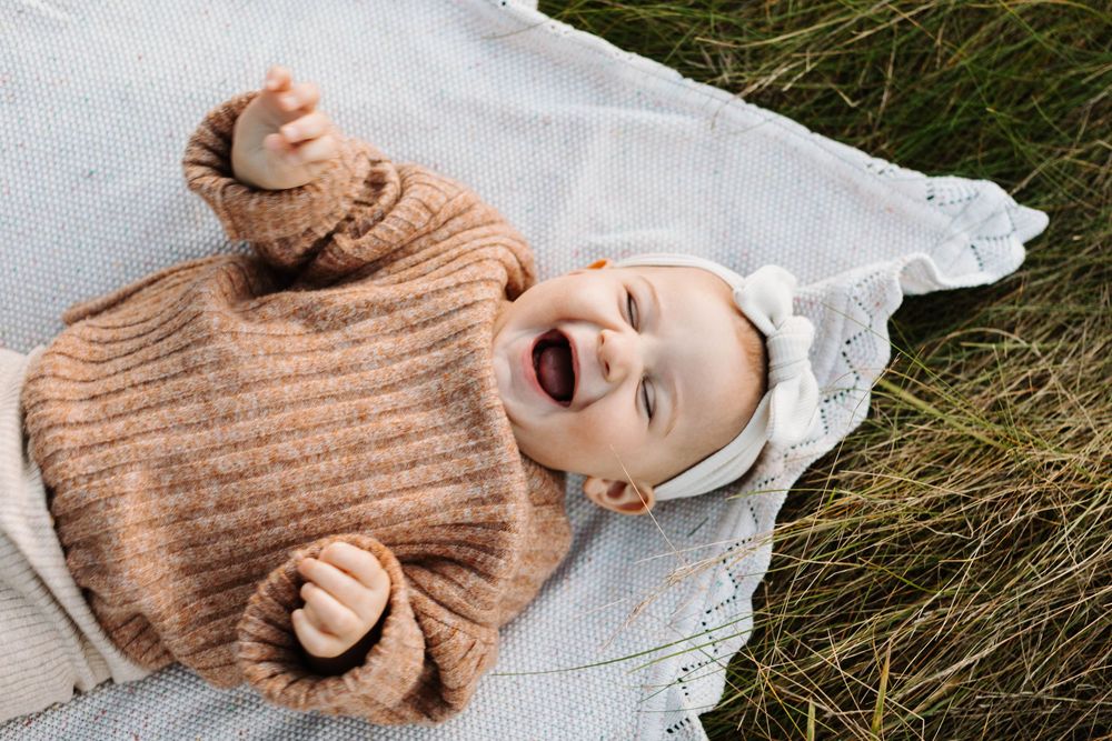 Baby in cozy brown sweater lying on white blanket in grass laughing joyfully in Guelph Ontario