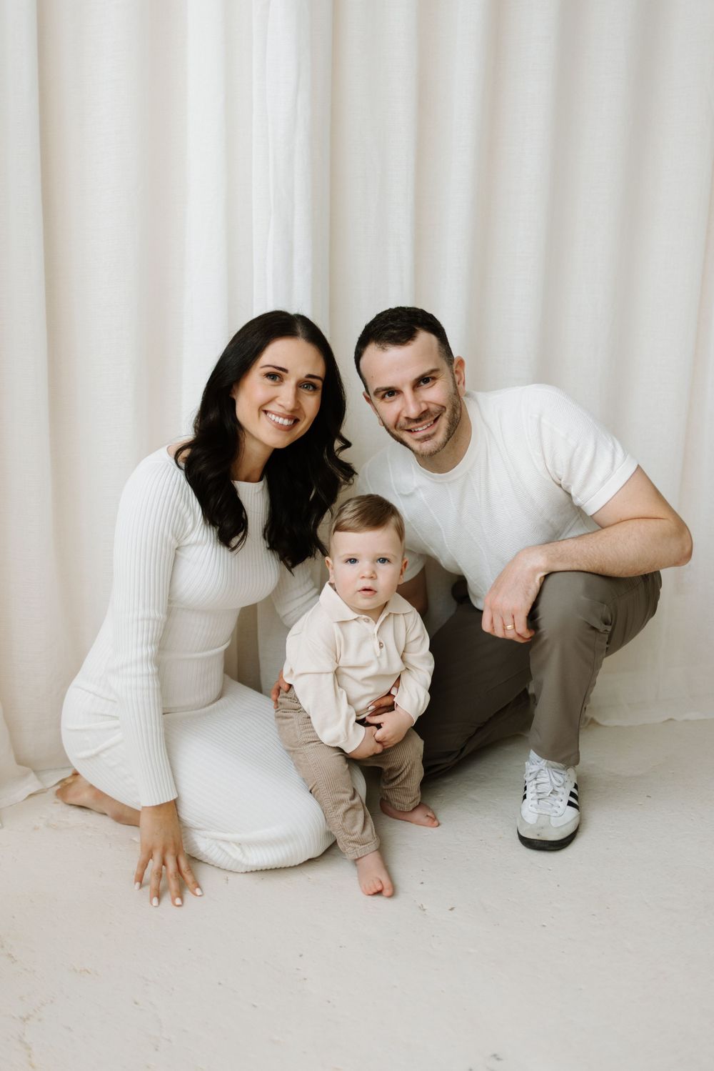A warm family studio portrait with three people wearing neutral colors against a cream curtain backdrop in Cambridge Ontario