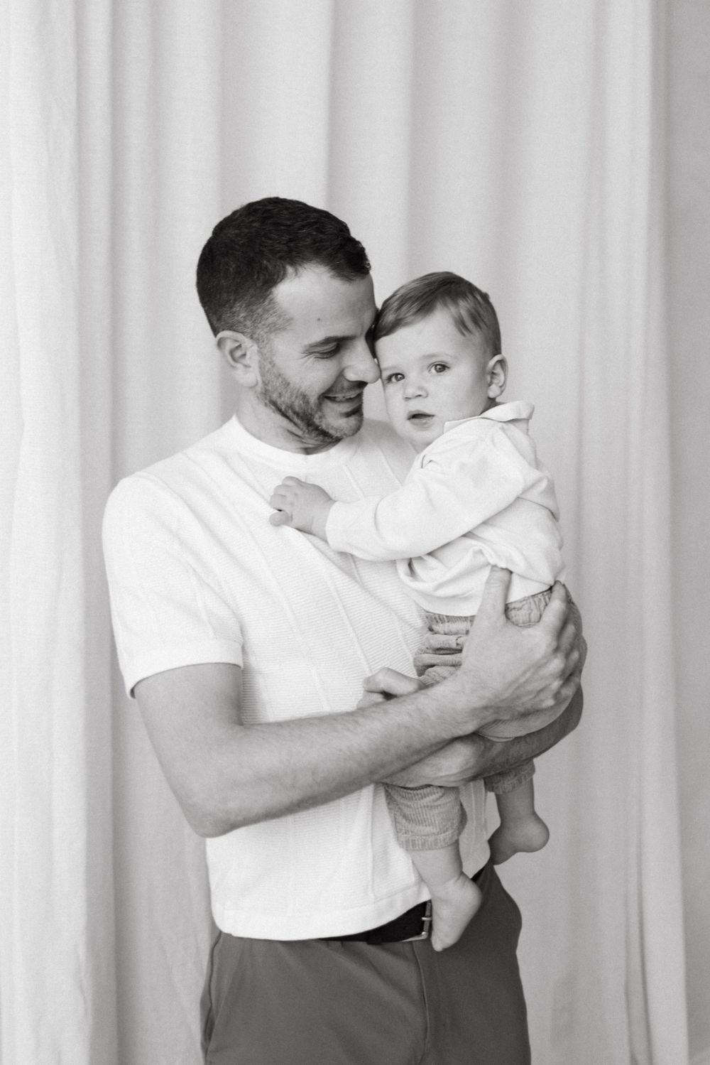 Intimate black and white portrait of a father holding and smiling at their young child against a light backdrop in Cambridge Ontario