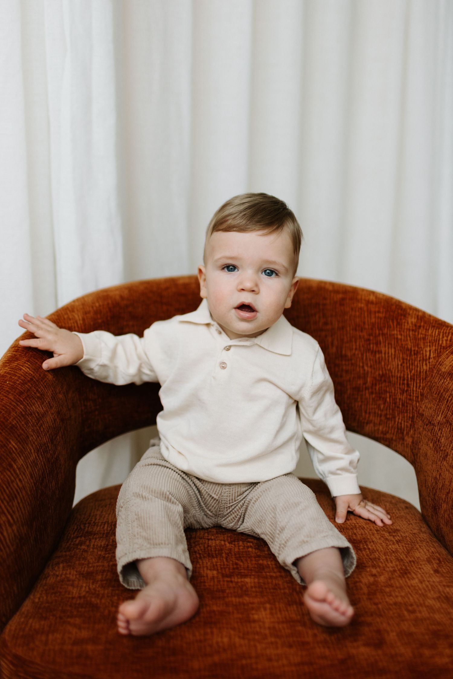 A toddler in a cream sweater and brown pants sits on a rustic orange armchair against a light curtain background in Cambridge Ontario