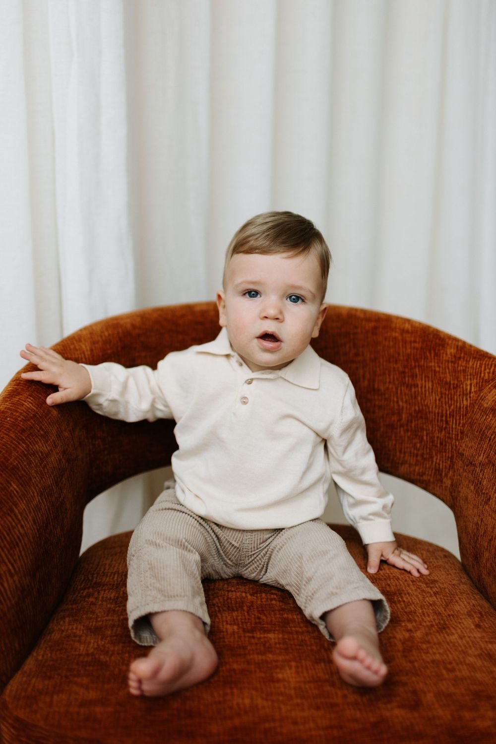 A toddler in a cream sweater and brown pants sits on a rustic orange armchair against a light curtain background in Cambridge Ontario