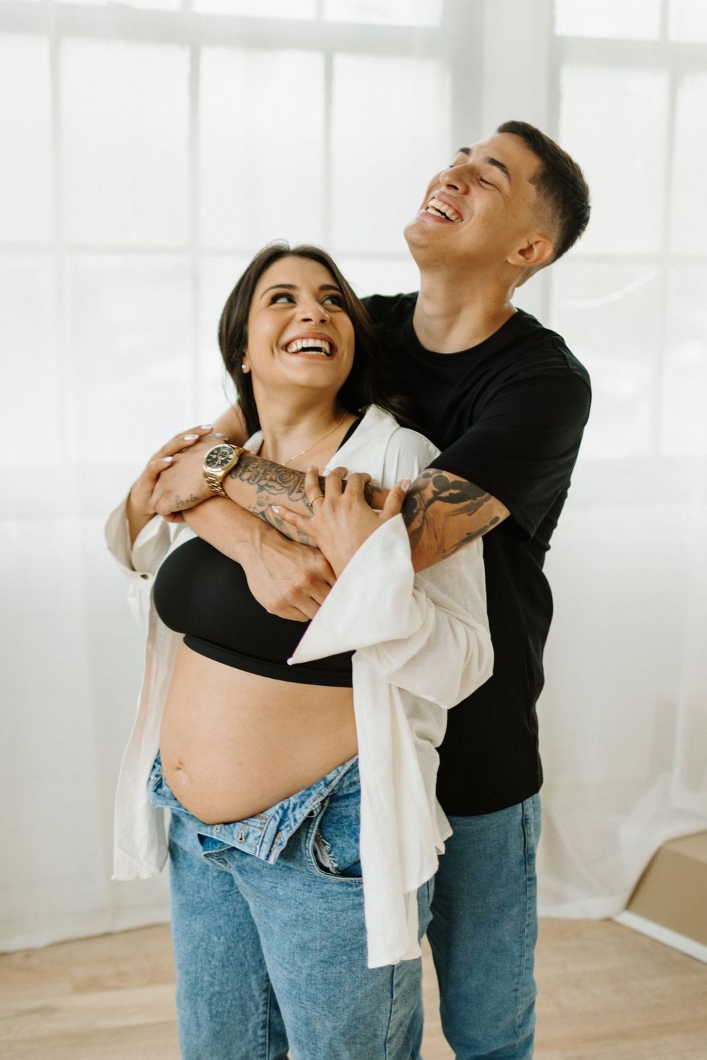 Young couple laughing and embracing in a bright indoor setting with white curtains and wooden floors in Cambridge Ontario