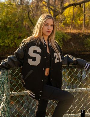 Fashion model in varsity jacket poses against autumn backdrop with chain link fence.