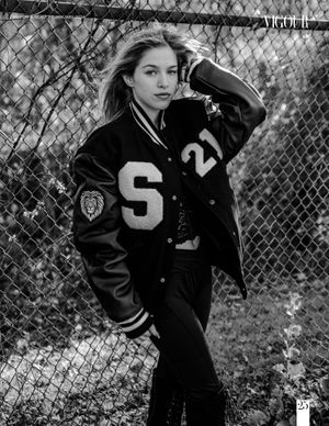 Black and white portrait of model wearing varsity letterman jacket against chain link fence.