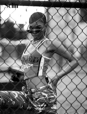 Black and white photo of someone posing in urban style clothing against a chain link fence background.