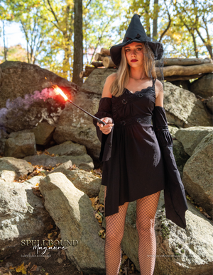 A witch in a black dress and hat casts magic with a glowing wand while posing dramatically on rocky terrain.