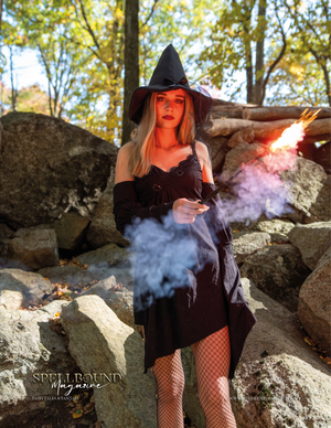 A witch in a black dress and hat poses among rocks while colorful smoke adds mystical atmosphere in a forest setting.