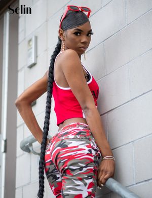 Athletic woman in red crop top and camo pants poses confidently against white wall with long braided hair.