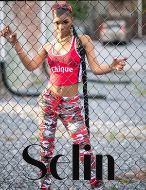A person poses in front of a chain link fence wearing a pink crop top and red camo pants with urban fashion style.