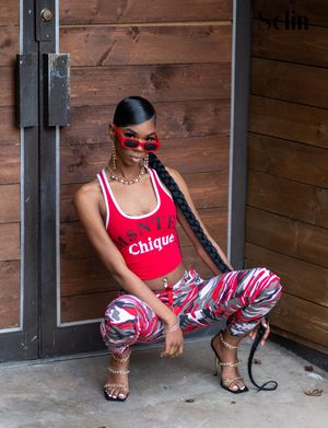 Fashion model in red tank top and patterned pants poses against wooden wall with sunglasses and gold accessories.