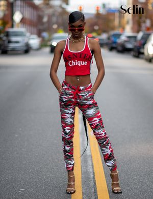 A person stands in the middle of a street wearing a red crop top and camo print pants with high heels.