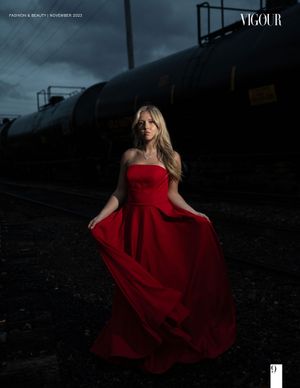Woman in flowing red gown poses dramatically near dark industrial train cars at dusk.