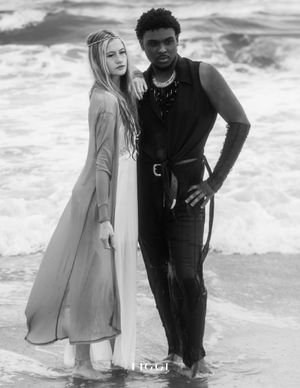 Artistic black and white fashion photo of two models posing together on a beach with waves in background.
