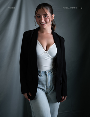 Fashion model poses in black blazer and white bodysuit against grey backdrop in studio portrait.