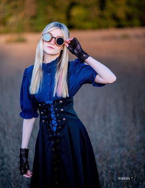A person in gothic-inspired fashion wearing a navy dress, corset, gloves and round sunglasses poses outdoors at sunset.
