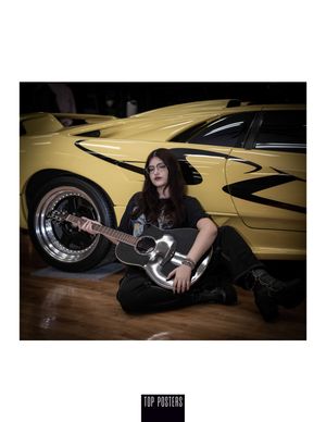 A musician in dark clothes sits on a wooden floor with an electric guitar next to a yellow sports car with custom details.