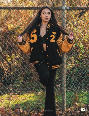 Model wearing varsity jacket poses against chain link fence in autumn setting.