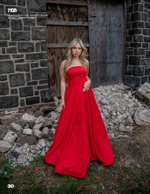 A person in an elegant red ball gown stands in front of a rustic stone wall and wooden door.