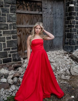 A person in a strapless red formal dress poses with hand in hair against weathered stone architecture.