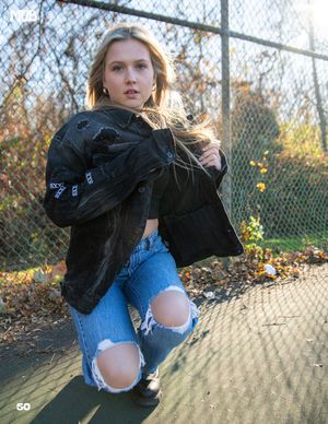 Fashion model wearing distressed denim and black jacket poses against chain link fence in autumn setting.