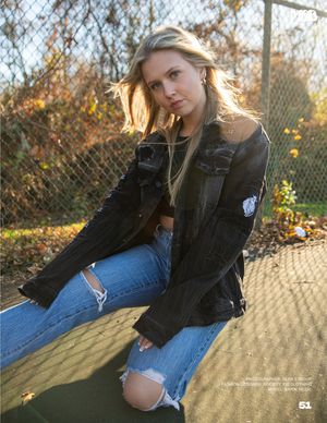 A person in distressed jeans and black denim jacket poses casually against fence at sunset.