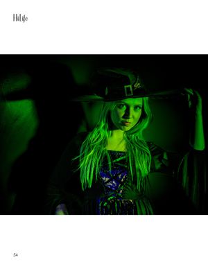 Figure in a witch hat illuminated by eerie green lighting against a dark background.