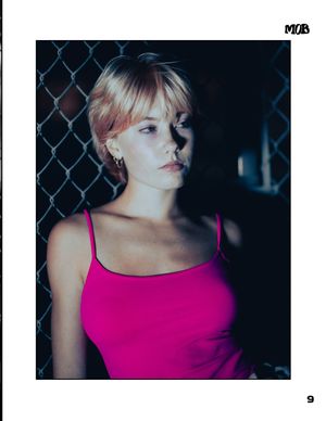 A person in a pink tank top poses against chain link fence at night.