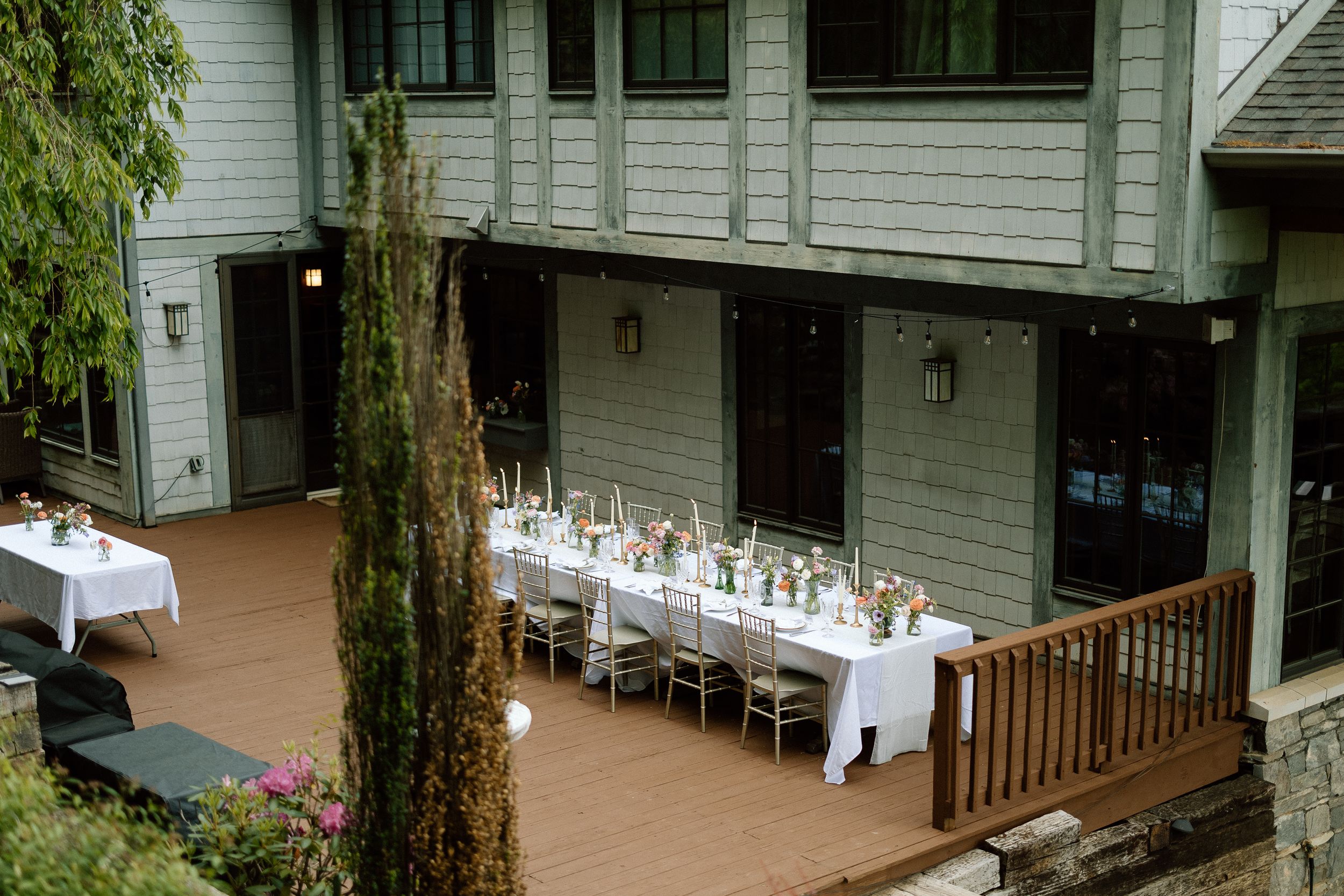 Long white tables set for an outdoor wedding reception on a wooden deck.