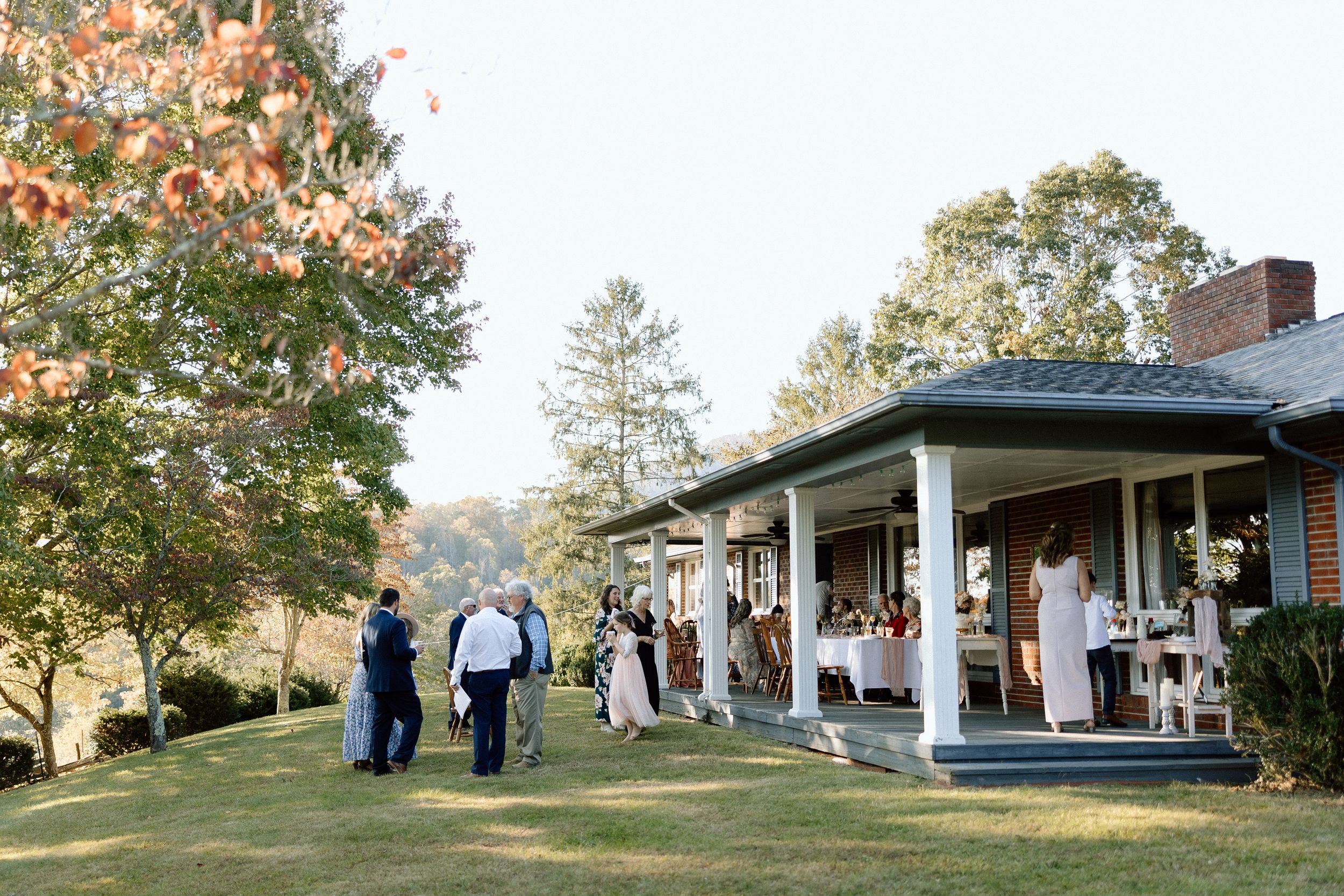 Guests gather outside a white farmhouse-style building for a rustic wedding celebration.