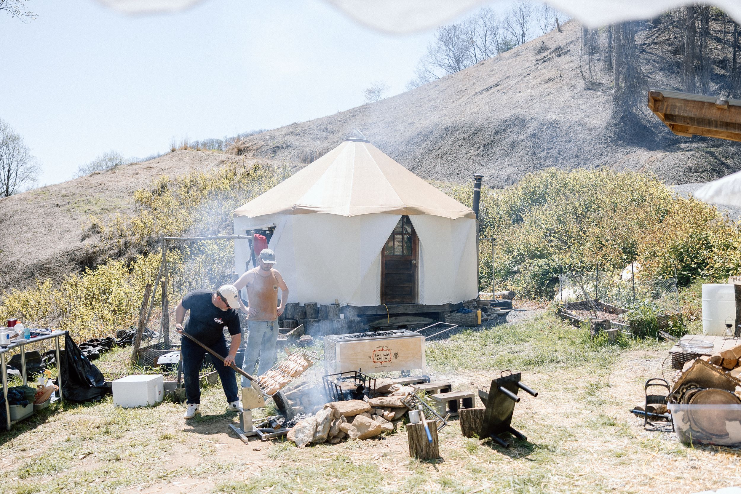 A rustic outdoor wedding setup with a canvas tent and campfire in a mountainous landscape.