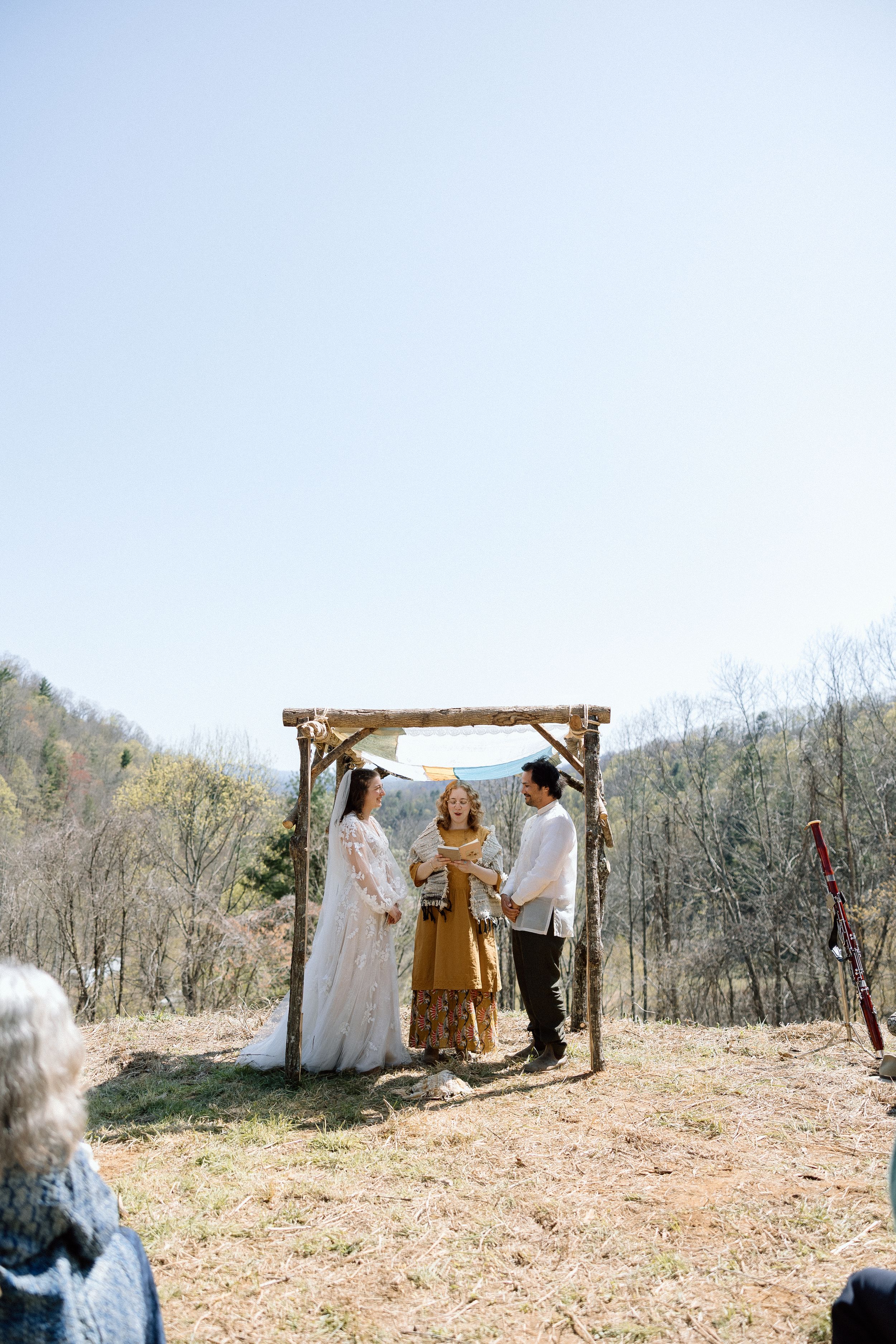 An intimate outdoor wedding ceremony beneath a wooden arch in a scenic mountain setting.