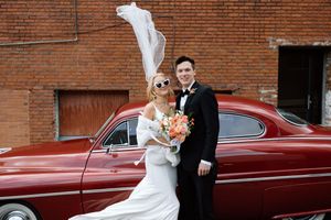 Elegant couple poses beside vintage red car with bride's veil flowing in the breeze.