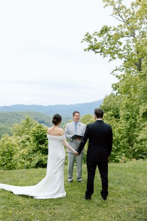 Intimate mountain wedding ceremony with couple and officiant against scenic forest backdrop.